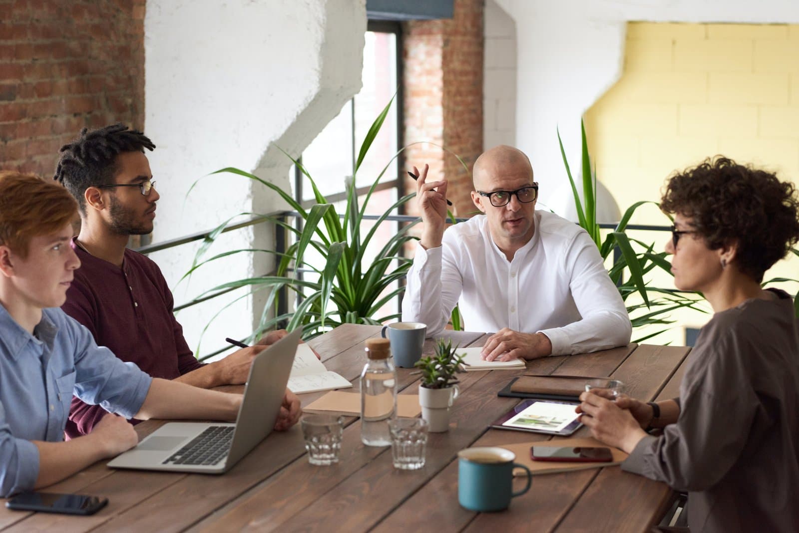 Four colleagues in discussion around a wooden table with a laptop, notebooks and plants