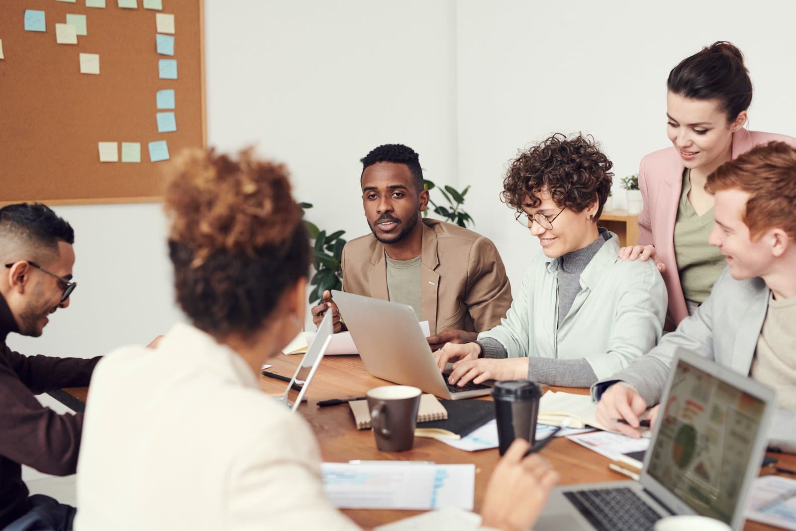 Five colleagues gathered around a meeting table with laptops and coffee, talking and smiling